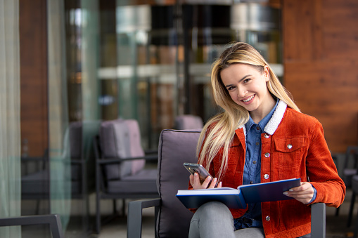 Facing the camera with a smile, a joyful student sits holding an open book and glancing at her phone. Her cheerful expression reflects the joy of learning in today's digital era. This image captures the seamless integration of technology and traditional study materials, perfect for illustrating modern study habits and educational enthusiasm.