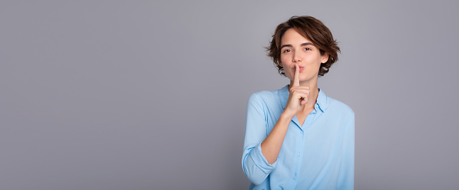 Banner photo of cute young brunette woman wearing smart casual clothes posing saying shush be quiet with finger on her lips shhh gesture looking camera isolated on gray background