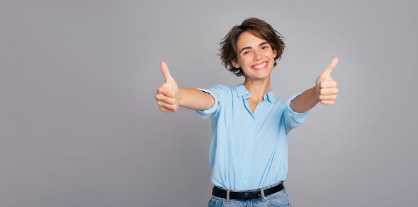 Banner photo of young smiling brunette woman with stylish hairstyle isolated on gray background showing or giving a thumbs up gesture on camera