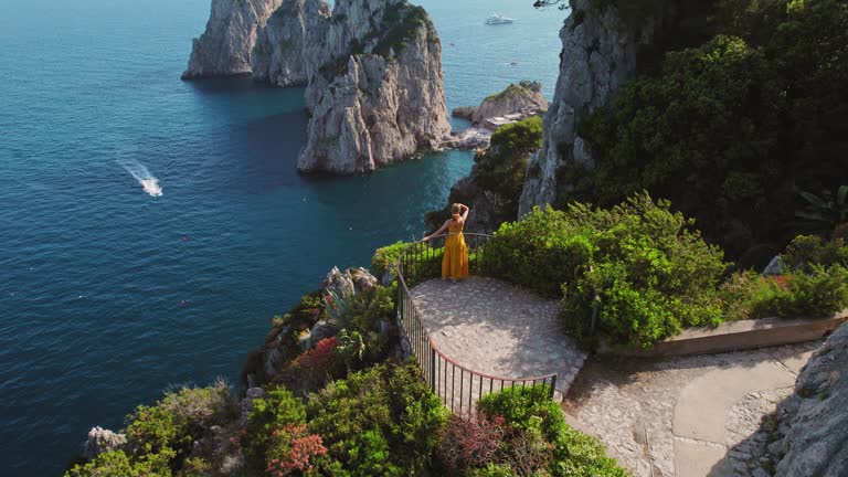 A woman in a yellow dress admires Capri scenic Faraglioni from a cliffside viewpoint, surrounded by the beauty of nature.