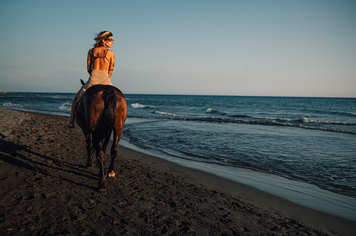 Back view of an tattooed equestrienne horseback riding at the beach at summertime at golden hour. Rear view of a young attractive tattooed lady horseback riding near sea water at summer. Copy space.