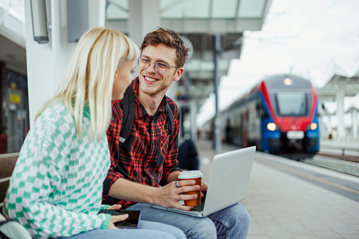 Portrait of a happy friends or a couple sitting on a bench at the railway station and drinking coffee and using a laptop. Man and woman waiting for a train to arrive and spending their time together.