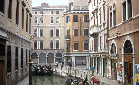 Morning street scene along the Venice, Italy canals includes man with delivery cart, parked gondolas, man reading paper. Photo taken August 28, 1967.