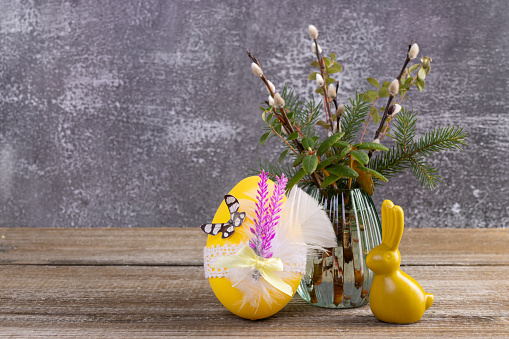 Easter composition with yellow bunny figurine and decorated egg near the vase with pussy willow branches and evergreen plants (labrador tea, bear berry, fir) on the wooden brown table.