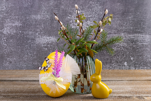 Easter composition with yellow bunny figurine and decorated egg near the vase with pussy willow branches and evergreen plants (labrador tea, bear berry, fir) on the wooden brown table.