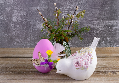 Easter composition on a brown rustic table: vase with bunch of branches from the wood (pussy willow, labrador tea, fir, bear berry), big pink decorated egg and a figurine of a bird.