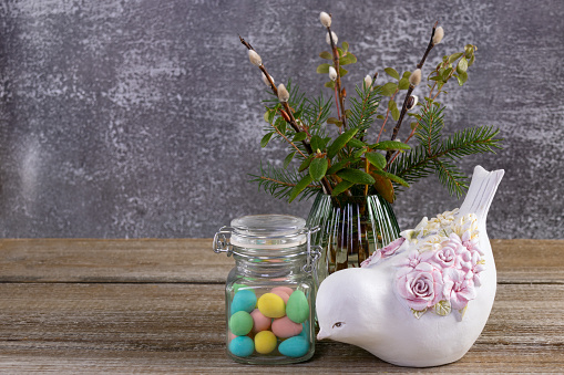 Easter composition on a brown rustic table: vase with bunch of branches from the wood (pussy willow, labrador tea, fir, bear berry), glass jar with colorful egg-shaped candies and a figurine of a bird.