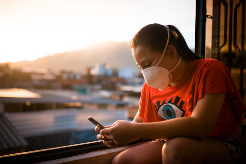 young woman wearing a face mask sitting by a window while using her mobile phone