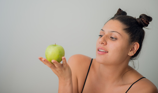 young latina woman with her hair tied back holding a green apple in her hands and looking at it with temptation