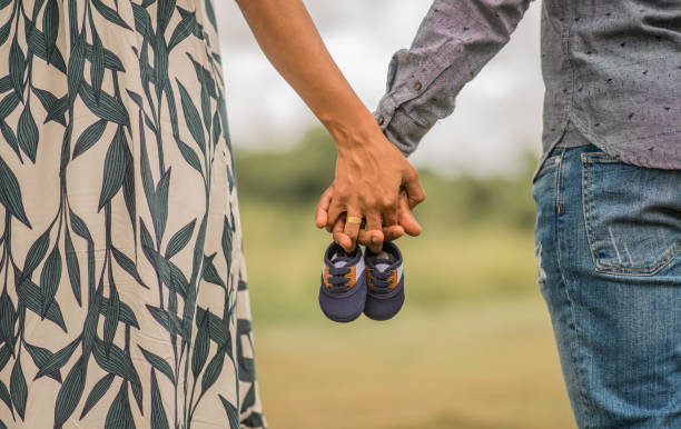 hands of two people outdoors clasped and holding two little baby shoes - pair couple mid adult happiness imagens e fotografias de stock