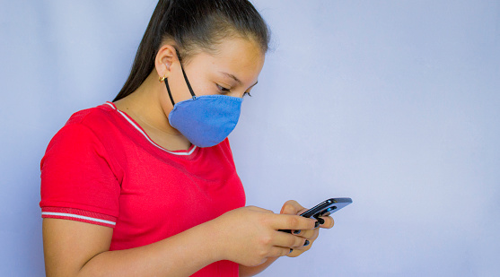 young woman wearing a mask on a white background while looking at her mobile phone in deep concentration.