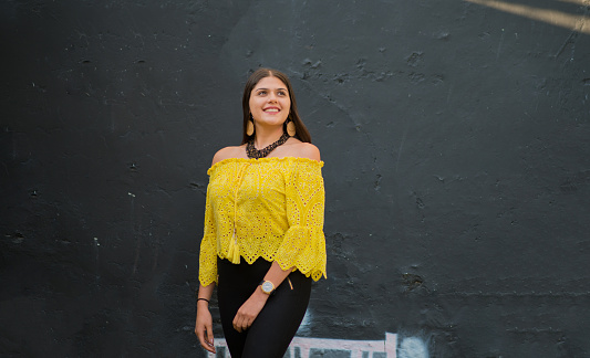 young latin woman smiling and looking to the side with her hands free and wearing colourful clothes against a background of a black wall.