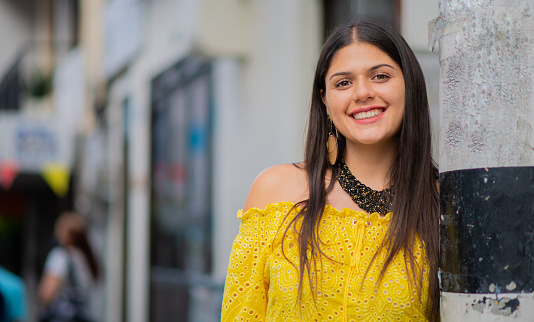 young latin woman with long hair smiling and looking at the camera while wearing a necklace and yellow blouse on a stroll through town
