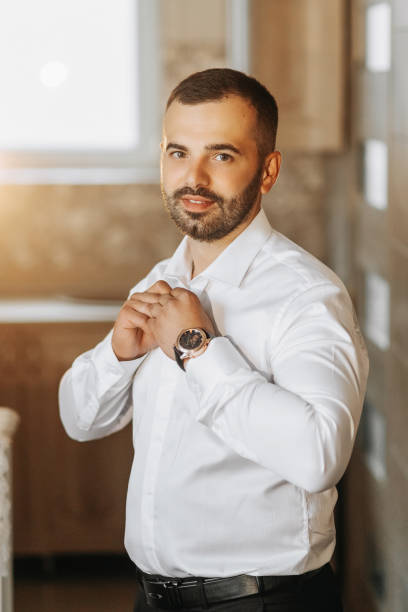 A young groom is buttoning up a white shirt while looking at the camera. The man is wearing a shirt. The groom is preparing for the wedding.