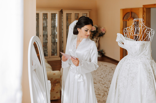 A beautiful bride reads a love letter from the groom. The bride in a white satin robe is standing near the mirror and the dress on the mannequin, reading a letter from her lover. Morning of the bride.