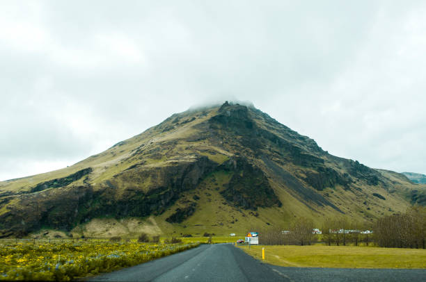 A road leading towards a beautiful volcanic mossy rocky mountain half-covered with clouds on an overcast day in Iceland.