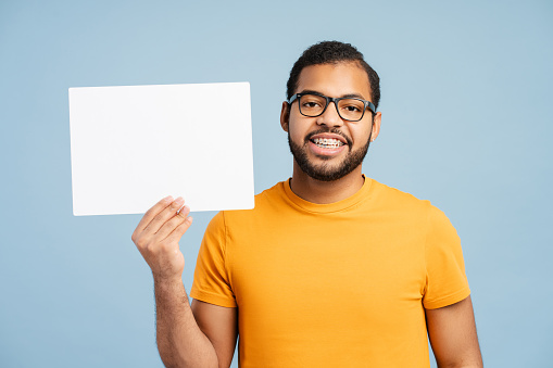 Portrait photo of a confident, smiling African American man with braces, wearing an eyewear, holding an empty poster while looking at the camera, isolated on a blue background. Mockup concept