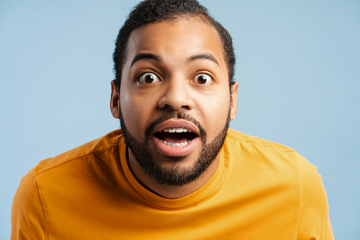 Portrait of a surprised African American student with dental braces, looking at the camera with his eyes and mouth wide open, isolated on a blue studio background