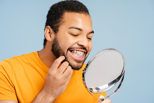 Portrait of an attractive African American man with orthodontic braces, examining his teeth while looking in a mirror, isolated on a blue background. Dental care concept