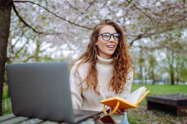 istock Positive student with laptop reading book while doing homework assignment on green meadow in campus park. 2085216130