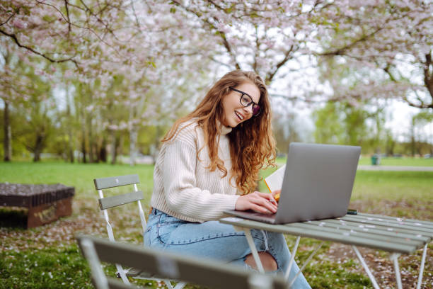istock Positive student with laptop reading book while doing homework assignment on green meadow in campus park. 2085216654