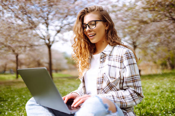 istock Positive student with laptop reading book while doing homework assignment on green meadow in campus park. 2085217174