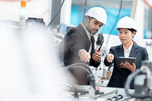 Industrial company employees are an American male engineer and an Asian female architect wearing suits, hard hats, using tablets, pointing at the circuit system of an automobile manufacturing machine.