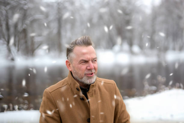 Middle-aged man with gray hair and a gray beard in front of a snowy winter landscape while it is snowing
