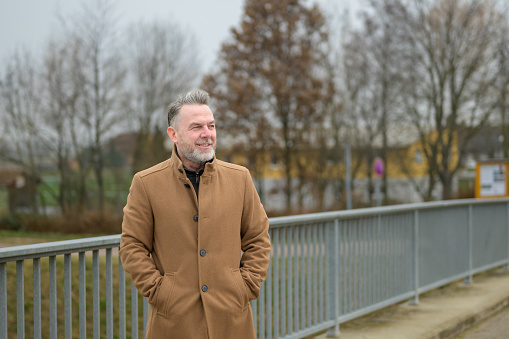 Sideview of amiddle aged man with gray hair and gray beard on a bridge in winter or spring with his hands in pockets