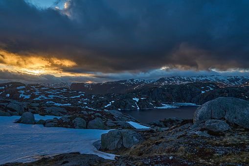 dramatic sunset over an idyllic mountain lake with ice on the water and snow covered peaks in the background along the Lysebotn road on a sunny day in Norway.