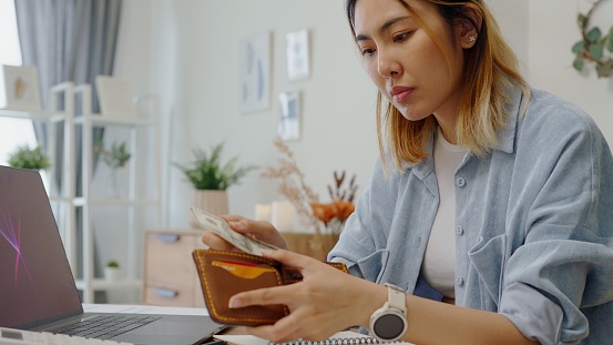 Struggle woman opening  wallet  with tiny US dollar having financial problems paychecks paper receipt bill statement ,calculating on smartphone for monthly expenses debt,planning budgets of monthly personal balance at home