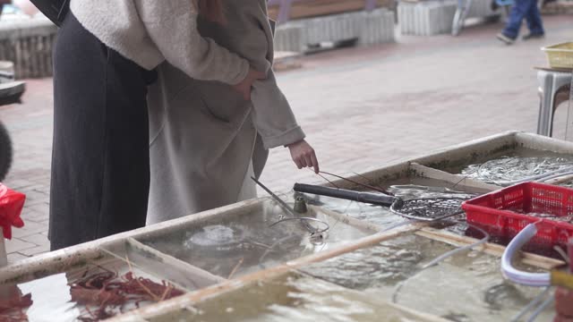 Two young Asian woman  are looking at a tank full of live seafood. They are dressed in a black coat and white jacket.