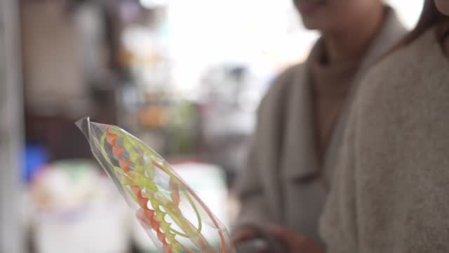 A young Asian woman with long dark hair is holding a clear plastic bag of colorful string candy. The woman is wearing a white shirt and the bag is held in her right hand.