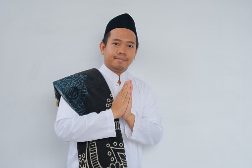 Young Asian Muslim man wearing white clothes and prayer rug on his shoulder, smiling and gesturing traditional greeting isolated against white background