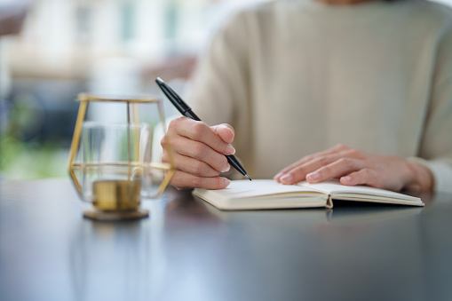 In the close-up shot, an Asian woman is seated at a table in her backyard, enjoying a moment of relaxation and me-time as she writes on a notepad in the morning.