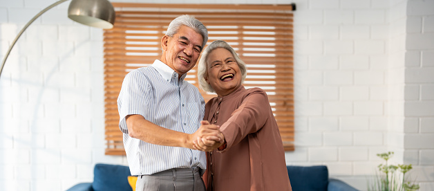 Portrait of Asian senior couple dance together in living room at home. Elderly older grandfather and grandmother smiling feeling in love and enjoy relationship and activity in house after retirement.