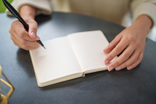 In the close-up shot, an Asian woman is seated at a table in her backyard, enjoying a moment of relaxation and me-time as she writes on a notepad in the morning.