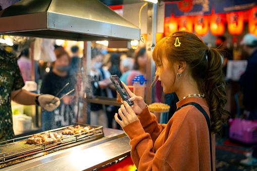 Young Asian woman using mobile phone taking picture of grilled beef steaks during travel street food night market in Taipei, Taiwan. Attractive girl enjoy and fun outdoor lifestyle nightlife shopping in the city on summer holiday vacation.