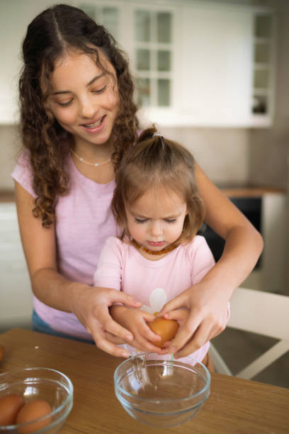 une adolescente et sa petite sœur cassent un œuf en préparant des biscuits à la maison - break eggs domestic kitchen breaking photos et images de collection