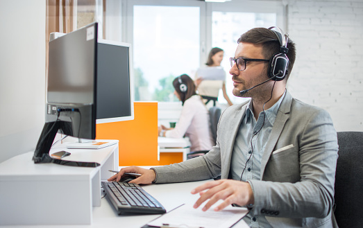 Confident male call center operator agent in headset with microphone consulting client online in customer support service office.