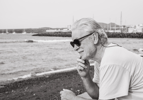 Black and white portrait of relaxed satisfied senior bearded man on sea beach at sunrise enjoying freedom, vacation or retirement concept