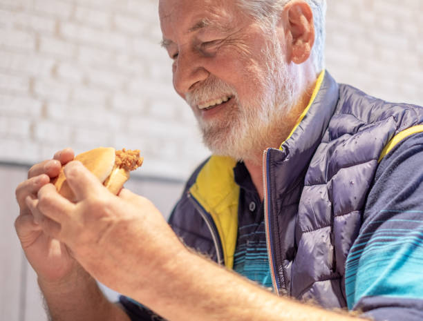 Bearded senior man sitting while happily eating a chicken sandwich in a fast food restaurant. Food and drink concept