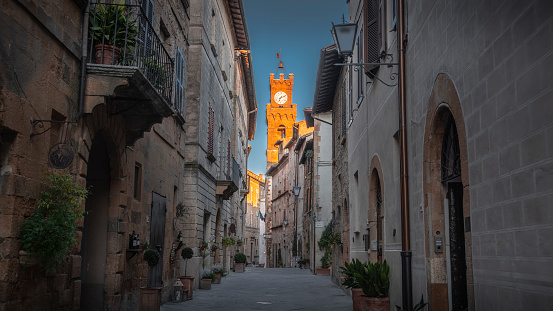 Narrow street in the old tuscan town Pienza in Italy. View on the clock tower of the town hall Palazzo Comunale illuminated in the first morning light. Situated in the province of Siena and in the famous region of Val d'Orcia Pienza is famous for its beautiful Renaissance architecture. Tuscany in Italy, Europe