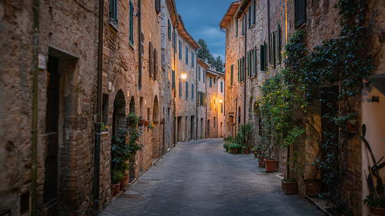 Cobblestone city street of old traditional Italian village San Quirico d’Orcia in Tuscany. The town is illuminated by the warm light of old street lanterns. Moody twilight sky at dawn. Tuscany in Italy, Europe