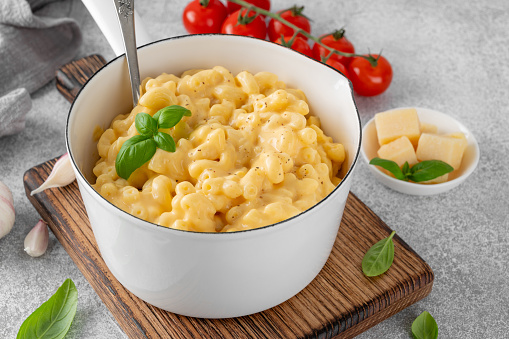 Mac and cheese in a white pot with fresh basil on top on a wooden board on a gray concrete background. American cuisine, comfort food. Copy space