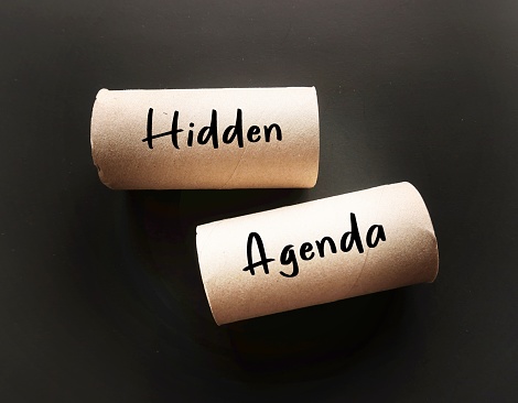 On black background, toilet paper roll with HIDDEN AGENDA, means secret reason for doing something, undisclosed plan or motive