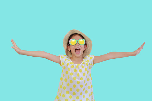 Cheerful Asian little child wearing a sunglasses and straw hat with open wide arms on cyan isolated background with clipping path. Summer and vacation concept
