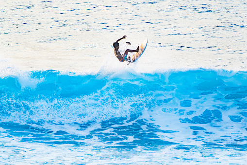 Bali, Uluwatu - A surfer is captured mid-motion, riding a wave against the backdrop of clear blue ocean water and white foam. The bright sunlight illuminates the scene, emphasizing the surfer’s body and surfboard.