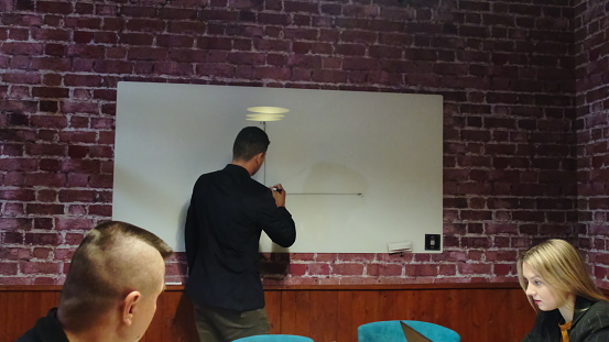 Businessman in business apparel draws graph on whiteboard during meeting in office. Co-workers analyze chart in contemporary boardroom