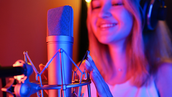 Young woman records a song into a studio microphone under neon LED lighting, blue and purple, in a recording studio
